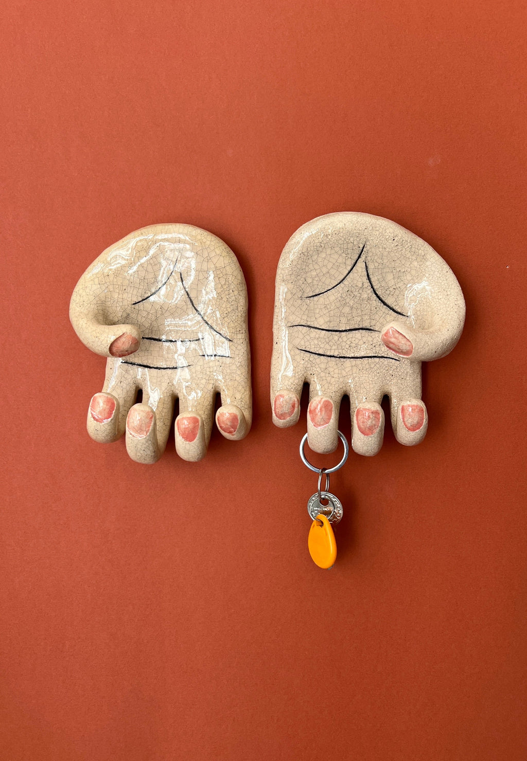 A pair of surrealist ceramic anatomical palm wall hangers (left and right hands) with visible hand-painted life lines mounted against a rich terracotta background.