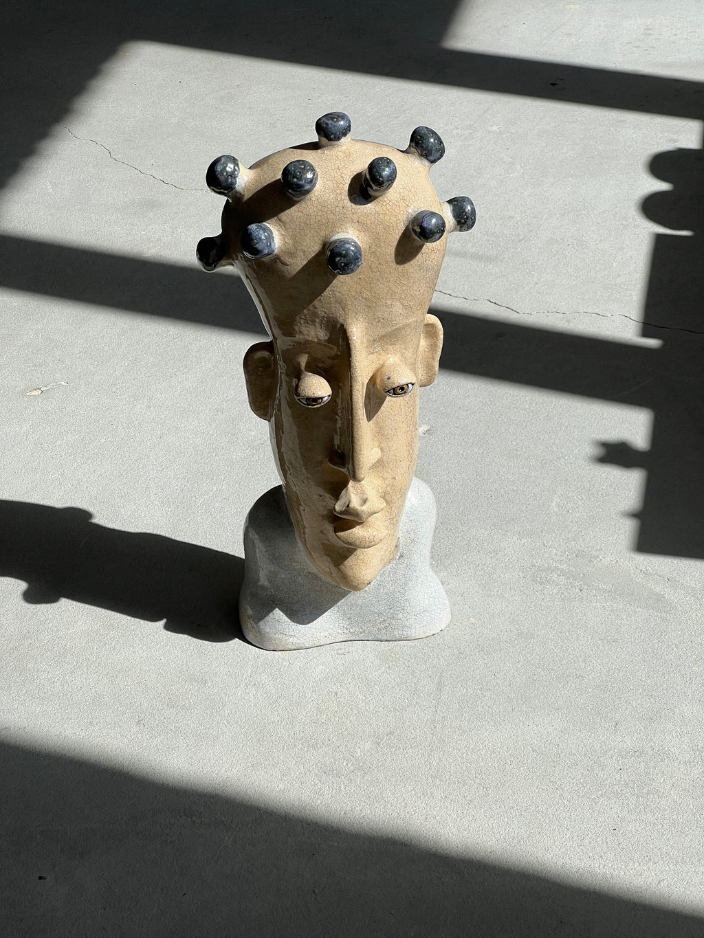 Top-angle view of a handmade ceramic bust sculpture resting on concrete, illuminated by harsh sunlight and shadows.
